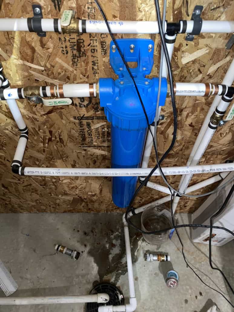 A whole-house water filter mounted on a bypass loop next to a tankless water heater, with rust showing on the steel fitting on the outlet side